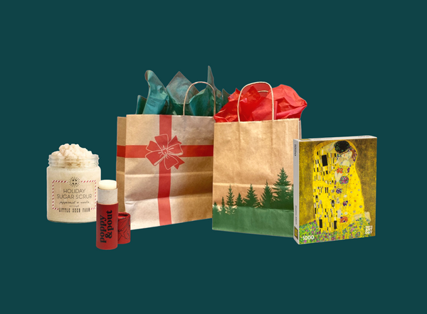 Shown from left to right: Peppermint and Vanilla Holiday Scrub, Cinnamint Lip Balm, Large gift bag with a red print bow and green tissue paper, a smaller gift bag with green printed trees and red tissue paper, and a puzzle featuring The Kiss by La Baiser